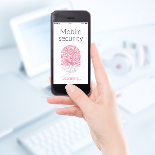 A woman with her phone in hand displaying a mobile security fingerprint scan. Her laptop and other devices are in the background. 